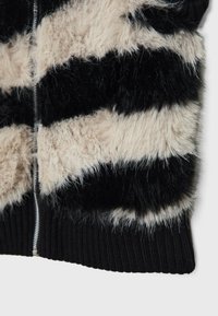 Veste douce et duveteuse rayée avec des bandes horizontales alternées en fourrure noire et beige et une fermeture éclair argentée visible sur un bord-côte noir.