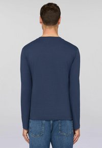 Mężczyzna odwrócony plecami, ubrany w gładką, granatową koszulę z długim rękawem i niebieskie dżinsy, stojący na jednolitym, jasnoszarym tle.