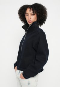 Ζακέτα σε σκούρο μπλε χρώμα με φερμουάρ, ψηλό γιακά, ελαστικά μανίκια και τελείωμα. Κατασκευασμένη από υφή υφάσματος, διαθέτει άνετη εφαρμογή και πλαϊνές τσέπες.