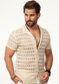 Beige gehäkeltes Hemd mit kurzen Ärmeln und Knopfleiste, das ein offenes Webmuster in Diamantform aufweist und eine lockere Passform hat. Mit Schmuck akzentuiert.