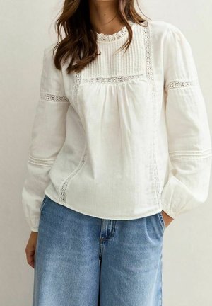 Žena v bílé blůze s dlouhými rukávy a krajkovými detaily, v modrých džínách, stojící s jednou rukou v kapse.