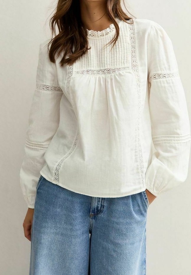 Femme portant un chemisier blanc à manches longues avec des détails en dentelle, associé à un jean en denim bleu, debout avec une main dans la poche.