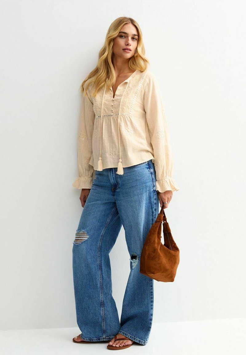 Cremefarbene Bluse mit langen Ärmeln, die mit Stickereien und Quasten verziert ist, kombiniert mit ausgefransten, weit geschnittenen blauen Jeans und einer braunen Wildledertasche.