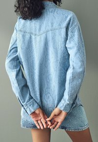 Camicia di jeans blu chiaro con un motivo testurizzato, maniche lunghe e orlo curvo, abbinata a pantaloni shorts in denim coordinati. Le mani sono dietro la schiena.