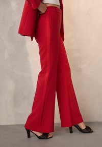 Punaiset räätälöidyt housut leveällä lahkeella, sileällä tekstuurilla ja sivusaumoilla. Yhdistetty mustiin korkokenkiin.