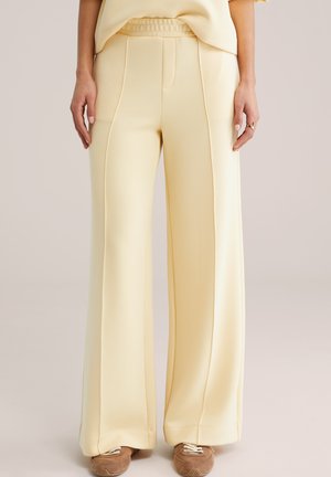 Kobieta ubrana w jasnopomarańczowe szerokie spodnie i pasujący top, w połączeniu z brązowymi zamszowymi trampkami, stojąca na neutralnym tle.