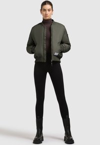 Olijfgroene bomberjack met ribgebreide boorden, voorrits en zijzakken, gecombineerd met zwarte leggings en enkelboots met stevige zolen.
