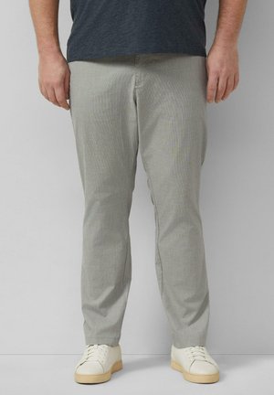 Homme portant un pantalon slim gris clair et des baskets blanches, debout devant un mur uni gris clair.