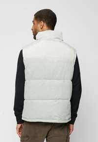 Chaleco acolchado ligero en corduroy beige y gris claro, que presenta un collar alto y un diseño acolchado con un acabado suave.
