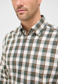 Camicia a quadri verde, marrone e bianco. Collo button-down, maniche lunghe e una tasca sul petto. Materiale in cotone con piccoli bottoni bianchi.