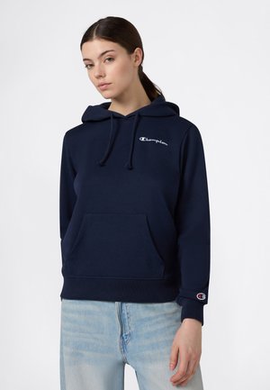 Vrouw draagt een marineblauwe Champion-hoodie met logo op de borst en manchetten, gecombineerd met lichtblauwe jeans, staand tegen een effen witte achtergrond.