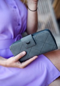 Gray quilted wallet made of textured material, featuring a snap closure and black logo, held in a hand against a purple dress.