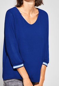 Blå lösblus med trekvartsärmar, prydd med vita accentsträck vid ärmsluten och en bred V-ringning. Slät tygstruktur.