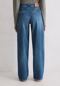 Achteraanzicht van een persoon in hoge taille, wijde blauwe jeans met achterste paspelzakken en een bruine leren merkpatch, staand tegen een witte muur.
