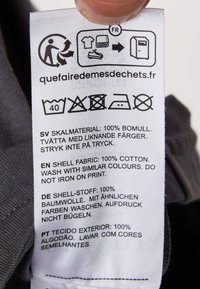 Hoitoetiketti, jossa on kankaan sisältö useilla kielillä. Materiaali: 100 % puuvillaa. Hoito-ohjeet: pese saman väristen vaatteiden kanssa, älä silitä kuviota.