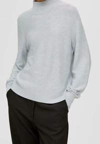 Hellgrauer Pullover aus weichem, strukturiertem Strick, mit hohem Kragen und lässiger Passform. Kombiniert mit dunklen Hosen, die einen entspannten Look zeigen.