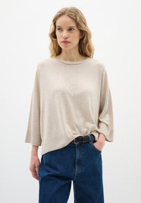 Beige kötött pulóver, csónaknyakú és széles ujjas, sötétkék farmernadrággal, magas derekú és fekete övvel párosítva.
