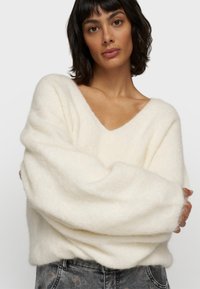 Weicher, cremefarbener, oversized Pullover mit V-Ausschnitt, voluminösen Ärmeln und einer flauschigen Textur. Kombiniert mit gemusterten, grauen Jeans.