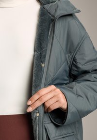 Person wearing a quilted gray jacket with faux fur lining, unzipping it to reveal a white top and brown skirt underneath.