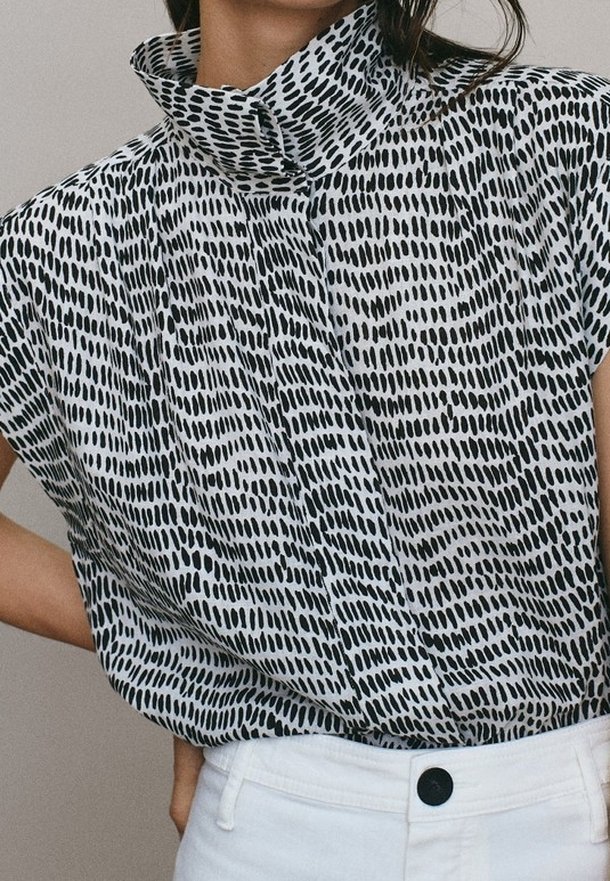Blouse à manches courtes à motifs noir et blanc avec col montant, rentrée dans un pantalon blanc taille haute avec un bouton noir.