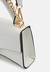 Sac à main rectangulaire blanc avec une texture crocodile, une bordure noire et une chaîne dorée fixée au rabat du sac.