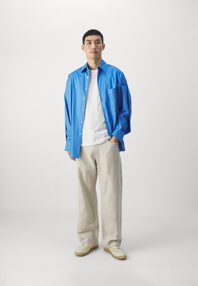 Camicia azzurro chiaro a maniche lunghe, t-shirt bianca, pantaloni beige e sneakers chiare, per un look rilassato e a strati.