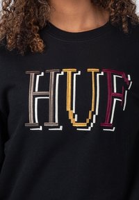 Felpa nera con lettere ricamate multicolori "HUF" in stili diversi, con tonalità di marrone, giallo, borgogna e bianco.