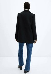 Abrigo de lana negro con un corte entallado, detalles en los hombros y un cuello con solapas, combinado con vaqueros acampanados azules y zapatos negros.