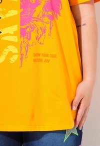 Camiseta de algodón amarilla con gráficos en rosa, que presenta un diseño audaz y el texto "MUESTRA TU VERDADERA NATURALEZA" en naranja. Corte casual.