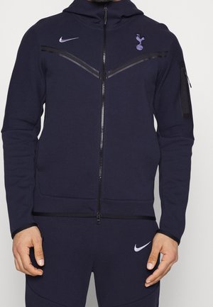 Mężczyzna ubrany w granatowy dres Nike z logo Tottenham Hotspur na piersi oraz kieszeniami na zamek na kurtce i spodniach.