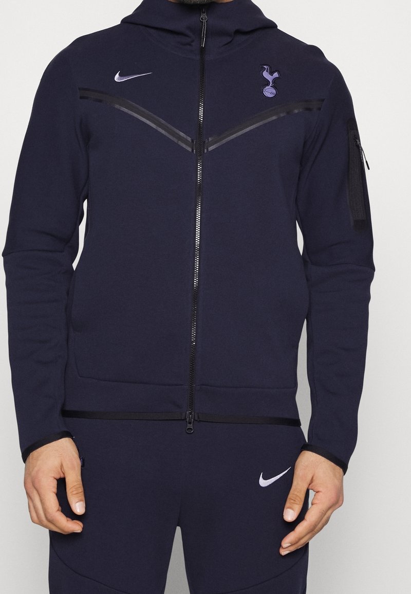 Homme portant un survêtement Nike bleu marine avec le logo Tottenham Hotspur sur la poitrine et des poches zippées sur la veste et le pantalon.