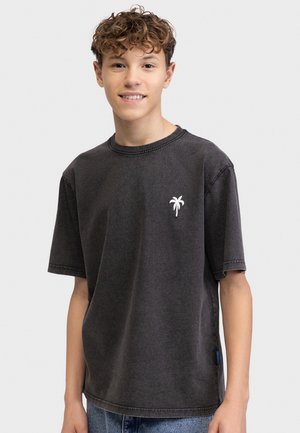 Tenårlig gutt med krøllete hår iført en falmet svart t-skjorte med en liten hvit palme-logo på brystet, stående mot en ensfarget bakgrunn.