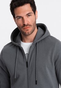 Graue Kapuzenjacke mit Reißverschluss und Kordelzügen. Trägt man über einem weißen Hemd mit schwarzem geometrischen Design. Weicher Stoff.