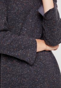 Tissu tricoté foncé moucheté avec des éclats rouges et blancs. Manches amples avec un détail de revers, texture lisse et silhouette décontractée.