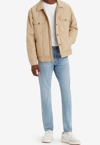 Beige vadderad jacka med två bröstfickor, knäppning med knappar och skjortkrage, i kombination med ljusblå jeans och vita sneakers.