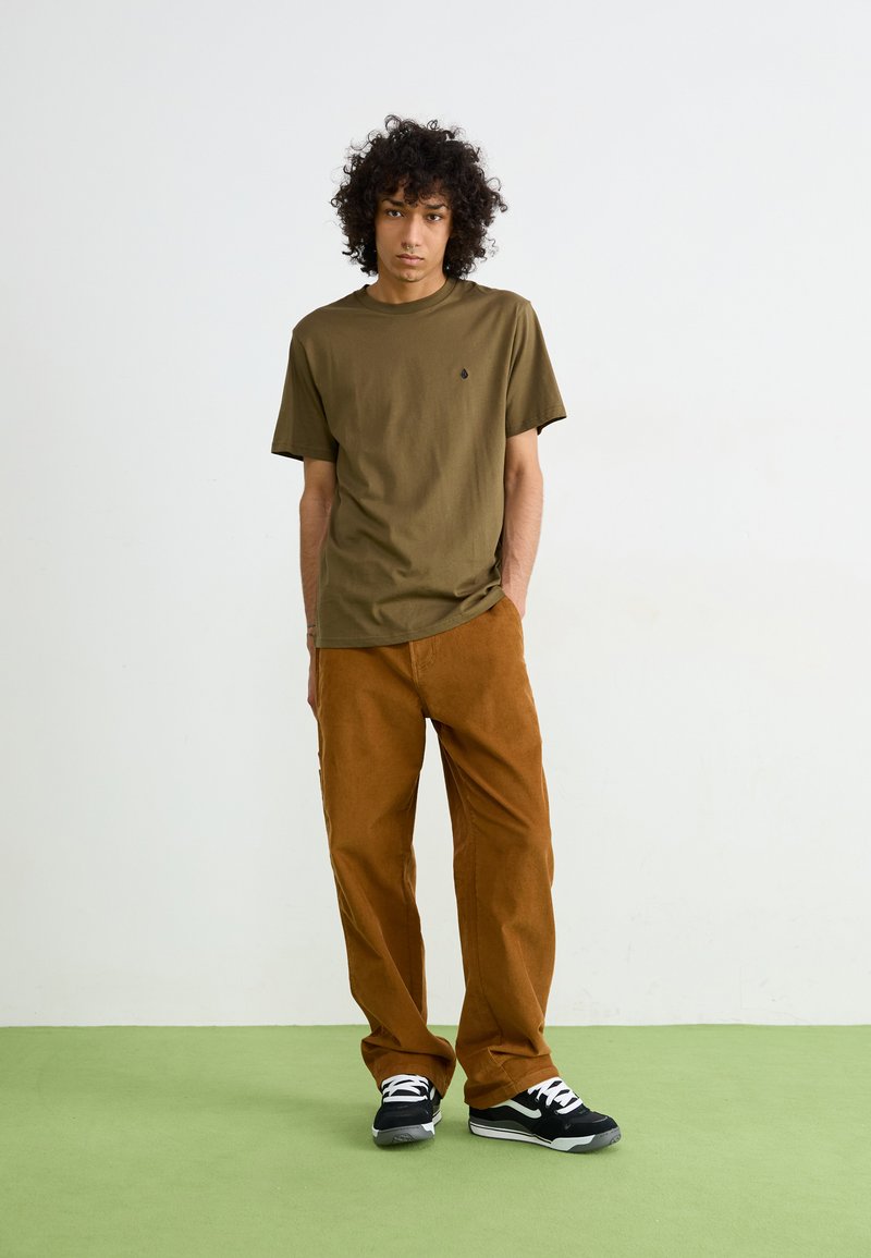 Olivgrünes Baumwoll-T-Shirt mit kurzen Ärmeln, kombiniert mit braunen Cordhosen. Schwarz-weiße Sneakers mit einer strukturierten Sohle.