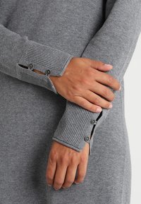 Grå strikket sweater med ribbede manchetter, der har knapdetaljer og sideslidser, som fremviser en blød tekstur og et simpelt design.
