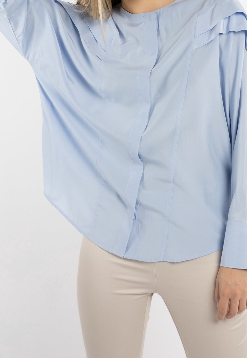 Una persona indossa una blusa azzurro chiaro con dettagli di pieghe unici, abbinata a pantaloni beige, e si trova di fronte a uno sfondo bianco semplice.