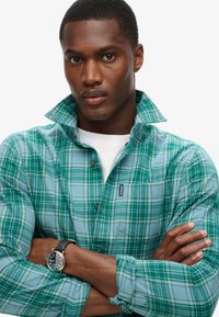 Camicia a quadri verde e blu con colletto chiuso, dotata di taschino sul petto e etichetta del marchio. Il modello indossa un orologio nero.