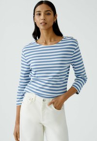 Blau-weiße gestreifte Langarmbluse mit rundem Ausschnitt und Knopfakzenten an einer Schulter, kombiniert mit weißen Jeans.