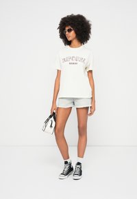 Lichtbeige t-shirt met "Rip Curl Hawaii" print, gecombineerd met lichtblauwe denim shorts. Zwarte hoge sneakers en een kleine zwart-witte handtas.