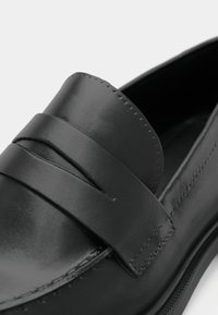 Mocassin en cuir noir avec une sangle cousue sur la partie supérieure et une finition lisse et polie.