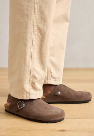 Barna velúr klumpák fém csattal és kerek gomb részlettel. Parafa talp és texturált gumi alap, bézs szűkített nadrággal párosítva.