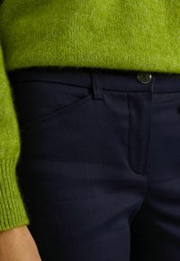 Manga y dobladillo de suéter verde de punto sobre pantalones azul oscuro con bolsillo delantero y cierre de botón, mostrados desde la cintura hasta la muñeca.