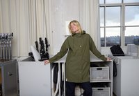 Alyvuogių žalumos neperšlampama striukė su gobtuvu, priekinių kišenių ir spaudžių užsegimais. Lygios tekstūros, iki kelių siekiantis dizainas. Neutrali fono aplinka su įranga.