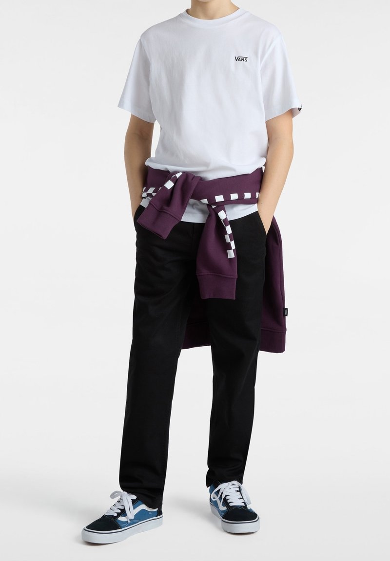 Barn som bär en vit Vans-t-shirt, svarta byxor, blå Vans-sneakers och en lila sweatshirt knuten runt midjan med ett rutmönster.