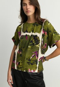 Haut floral vert à manches courtes présentant un motif abstrait blanc, un tissu texturé et une coupe ample, associé à un pantalon noir.