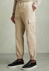 Joggers cargo beige con puños elásticos y cinturilla ajustable, que cuentan con bolsillos laterales y un bolsillo con solapa en el muslo. Combinados con zapatillas negras.