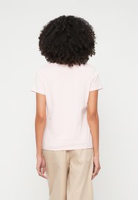 Lühikeste varrukatega, heleda roosa värvi t-särk pehmest puuvillast, ümar kaelus ja vabalt langev lõige, kantud beežide pükstega. Näidatud tagantvaates.