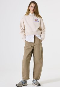 Lichtbeige cropped sweatshirt met print, gelaagd over een witte shirt. Ruimvallende khaki broek en grijze sneakers met blauwe accenten.
